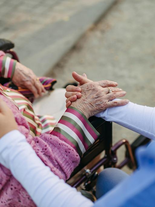 Eine Altenpflegerin hält die Hand einer alten Dame, die im Rollstuhl vor ihr sitzt.