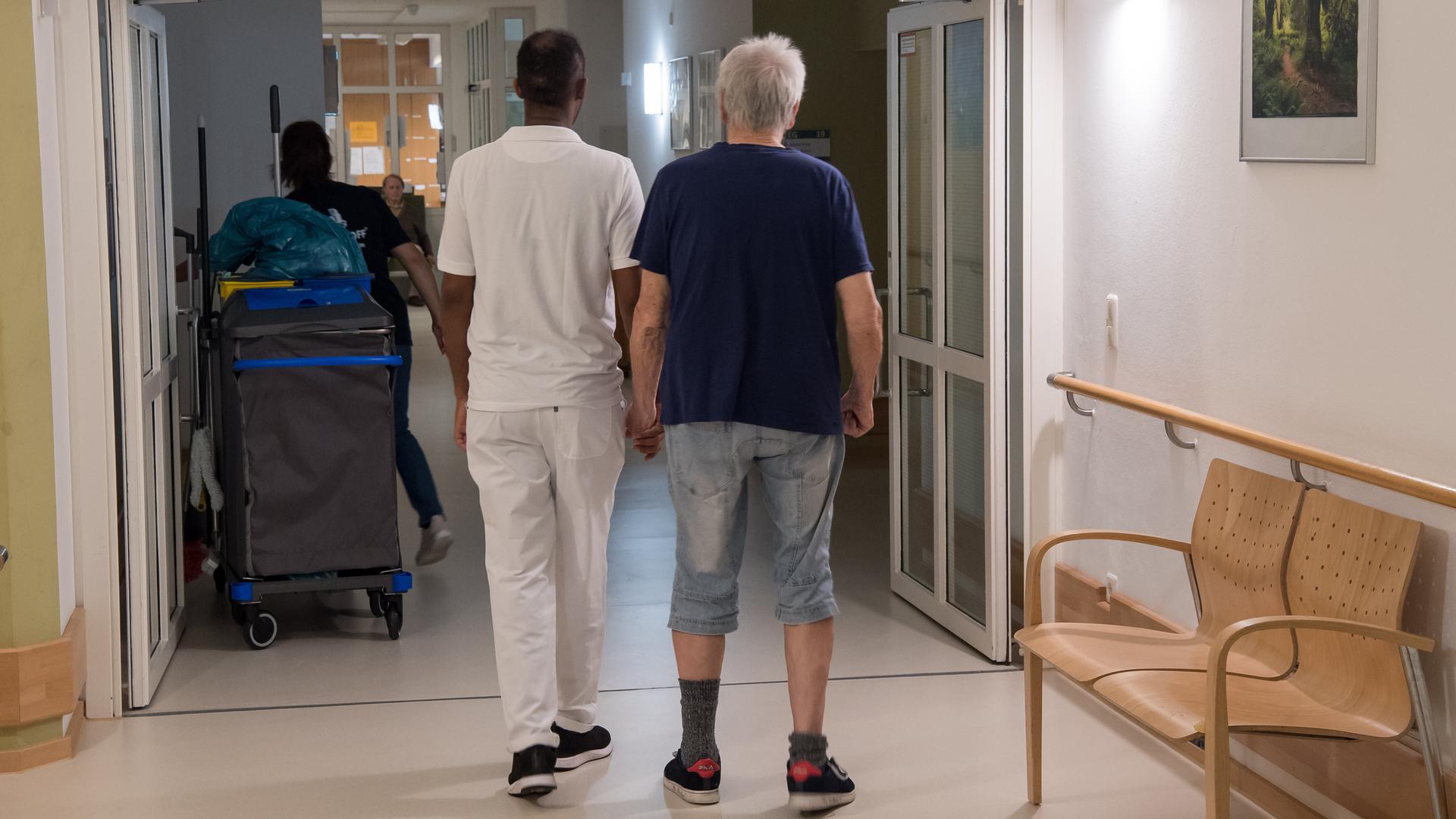 Ein pflegebedürftiger Mann (r) läuf mit einem Pfleger über den Flur.  