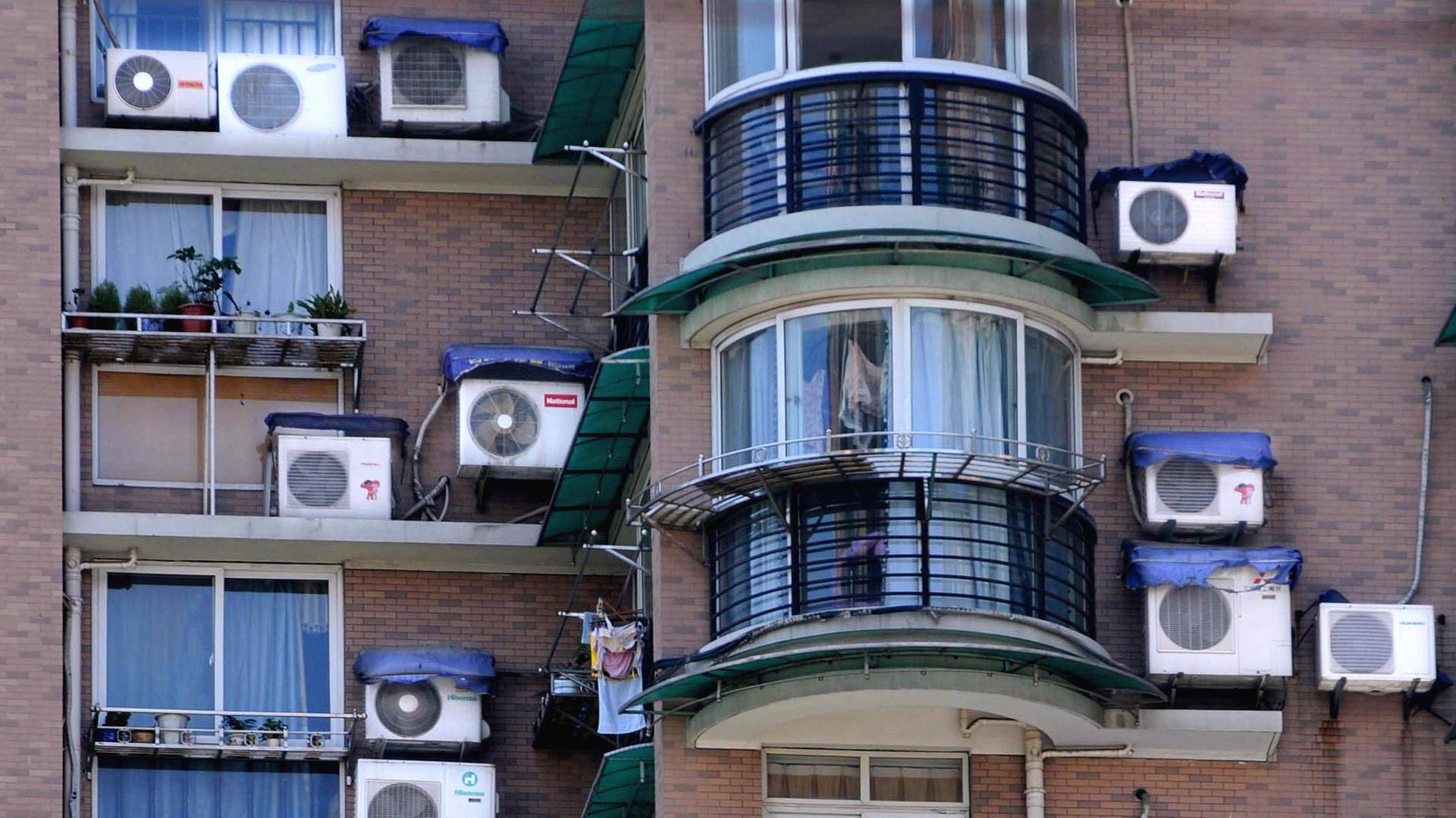 Klimaanlagen an einer Hauswand neben Fenstern und Balkonen im Sommer.