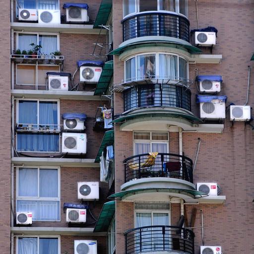 Klimaanlagen an einer Hauswand neben Fenstern und Balkonen im Sommer.