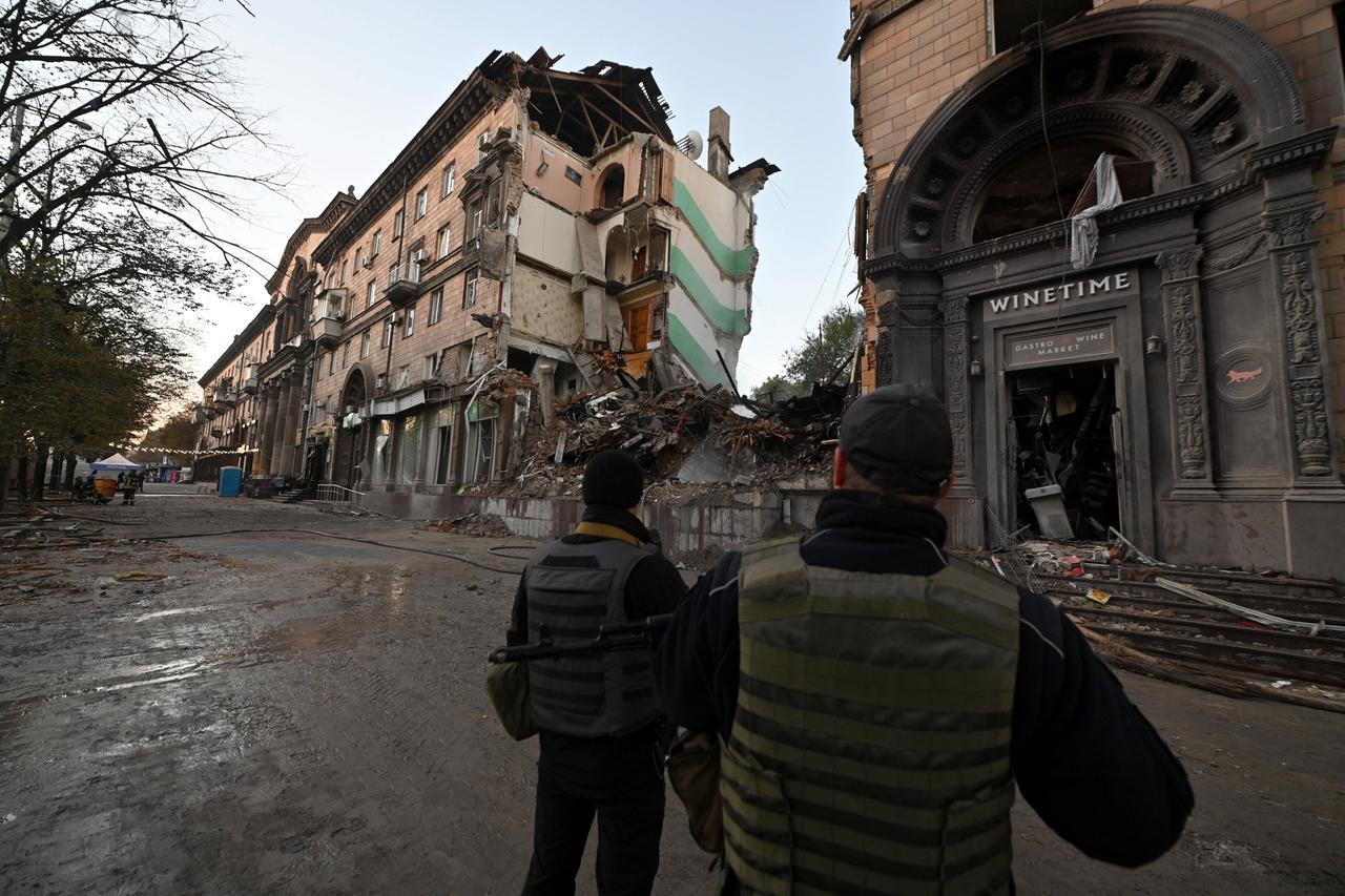 Zwei Männer in schusssicheren Westen stehen in Saporischschja auf der Straße und betrachten ein zerstörtes Wohnhaus.