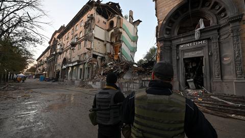 Zwei Männer in schusssicheren Westen stehen in Saporischschja auf der Straße und betrachten ein zerstörtes Wohnhaus.