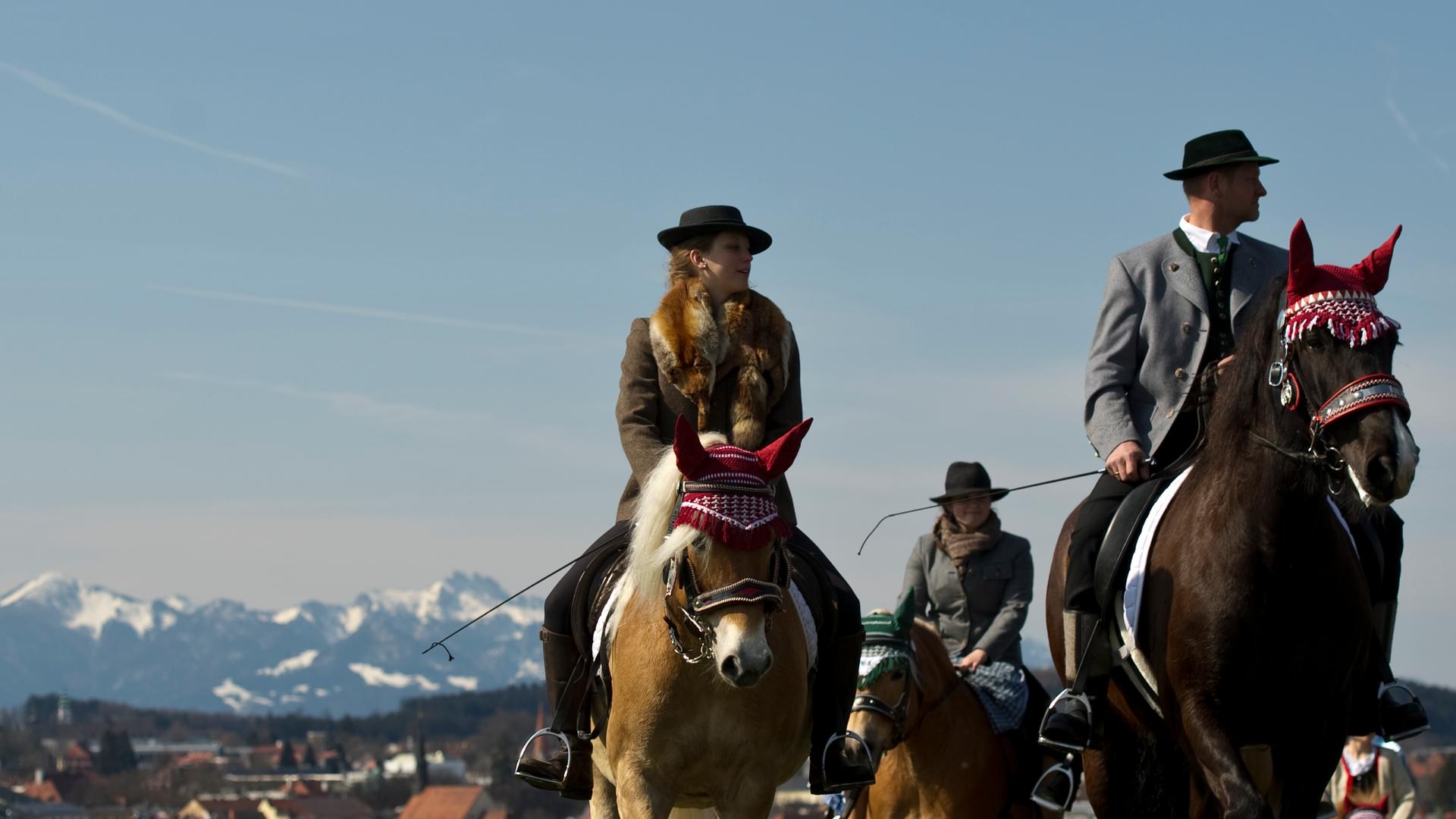 Zwei Frauen und ein Mann in Tracht reiten mit ihren geschmückten Pferden über eine Alm.