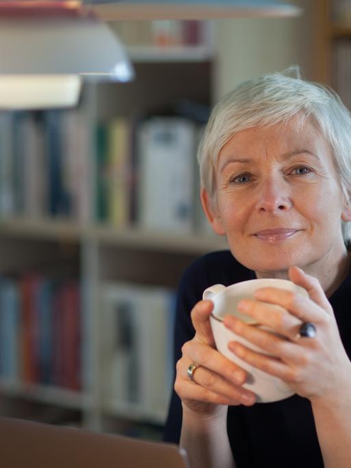 Portrait von Dörte Hansen, sie hät eine Tasse in beiden Händen, im HIntergrund ein Bücherregal Portrait von Dörte Hansen, sie hät eine Tasse in beiden Händen, im HIntergrund ein Bücherregal