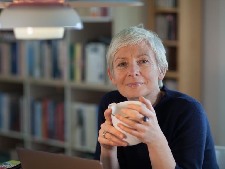 Portrait von Dörte Hansen, sie hät eine Tasse in beiden Händen, im HIntergrund ein Bücherregal Portrait von Dörte Hansen, sie hät eine Tasse in beiden Händen, im HIntergrund ein Bücherregal