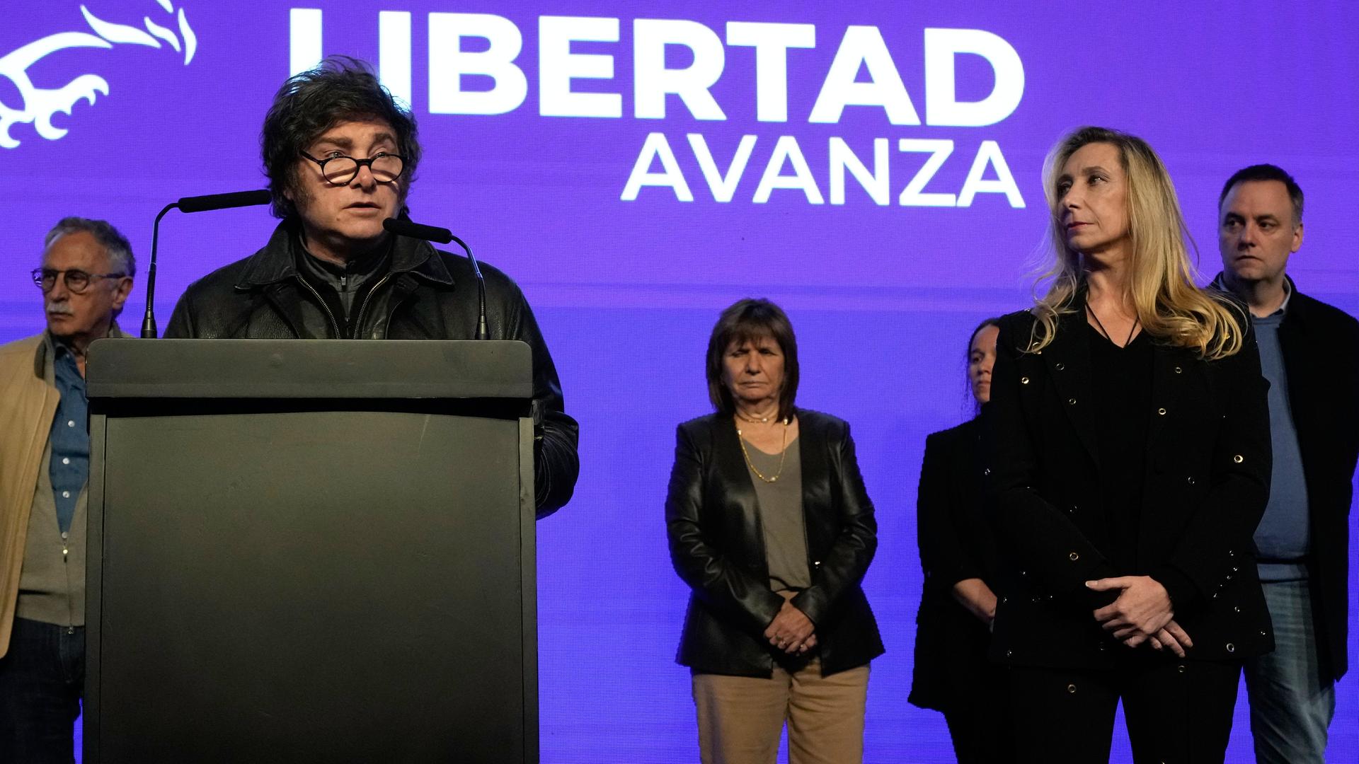 Javier Milei steht an einem Pult, weitere Personen sind neben und hinter ihm. Im Hintergrund an der lilafarbenen Wand befindet sich der Name seiner Partei in weißer Schrift: La Libertad Avanza. Javier Milei steht an einem Pult, weitere Personen sind neben und hinter ihm. Im Hintergrund an der lilafarbenen Wand befindet sich der Name seiner Partei in weißer Schrift: La Libertad Avanza.