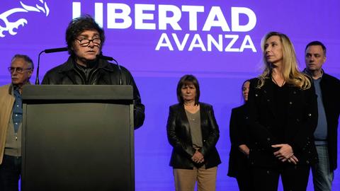 Javier Milei steht an einem Pult, weitere Personen sind neben und hinter ihm. Im Hintergrund an der lilafarbenen Wand befindet sich der Name seiner Partei in weißer Schrift: La Libertad Avanza.