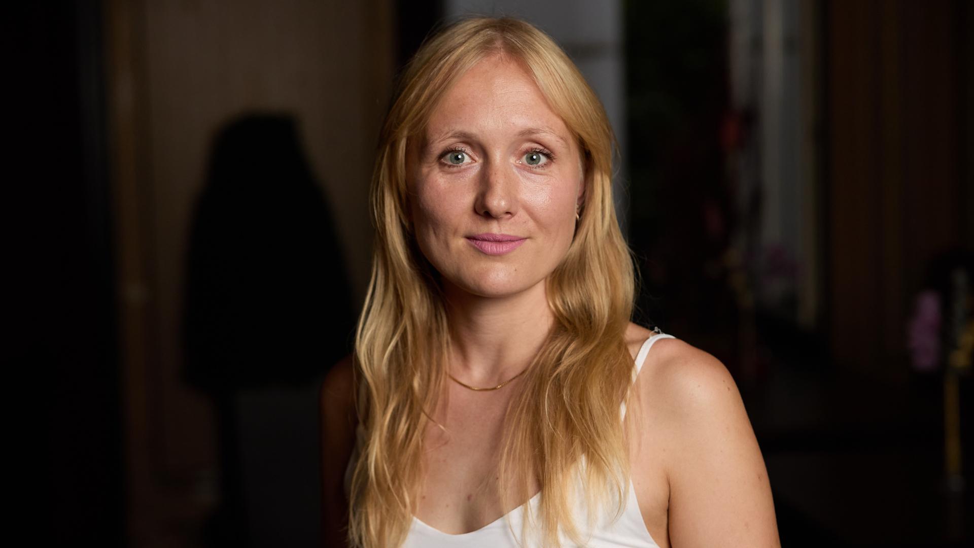 Die Autorin Ruth-Maria Thomas steht in einem weißen Oberteil vor dunklem Hintergrund.