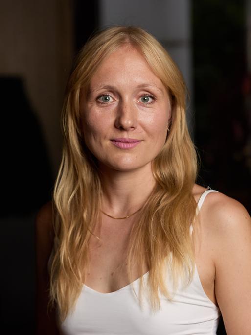 Die Autorin Ruth-Maria Thomas steht in einem weißen Oberteil vor dunklem Hintergrund. Die Autorin Ruth-Maria Thomas steht in einem weißen Oberteil vor dunklem Hintergrund.