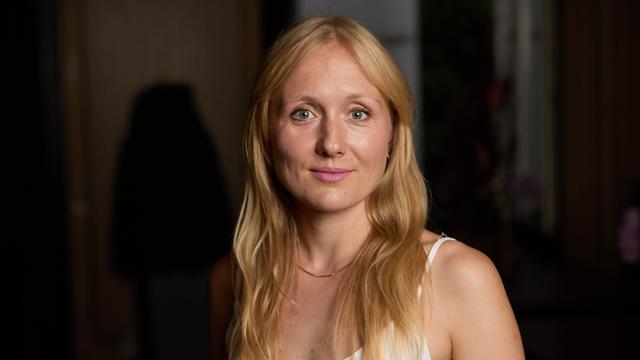 Die Autorin Ruth-Maria Thomas steht in einem weißen Oberteil vor dunklem Hintergrund.