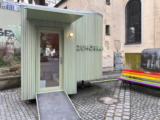 An einem oval geformten lindgrünen Holzbau neben der kleinen Kirche am Alten Südfriedhof in München steht "Zuhörraum".