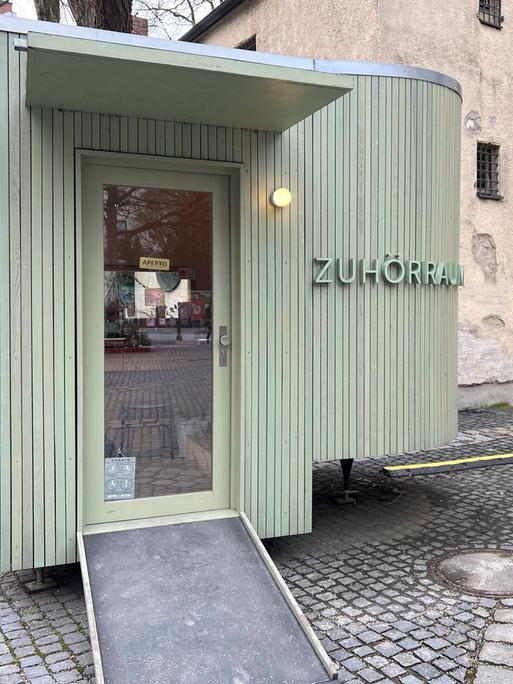 An einem oval geformten lindgrünen Holzbau neben der kleinen Kirche am Alten Südfriedhof in München steht "Zuhörraum".