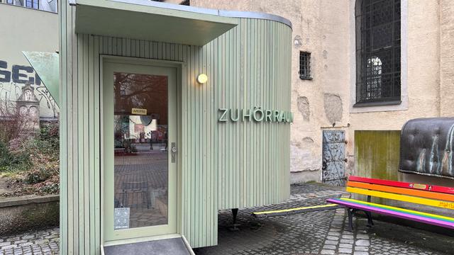An einem oval geformten lindgrünen Holzbau neben der kleinen Kirche am Alten Südfriedhof in München steht "Zuhörraum".