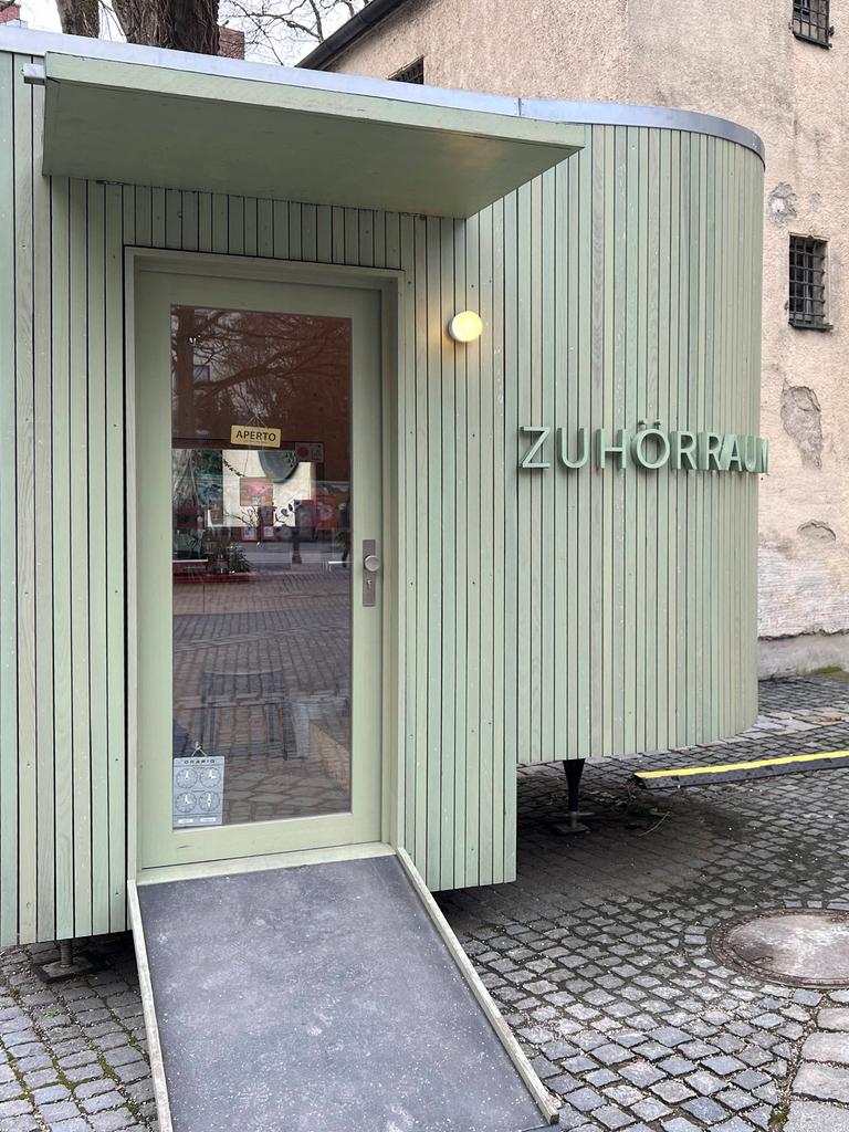 An einem oval geformten lindgrünen Holzbau neben der kleinen Kirche am Alten Südfriedhof in München steht "Zuhörraum".