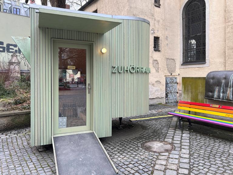An einem oval geformten lindgrünen Holzbau neben der kleinen Kirche am Alten Südfriedhof in München steht "Zuhörraum".