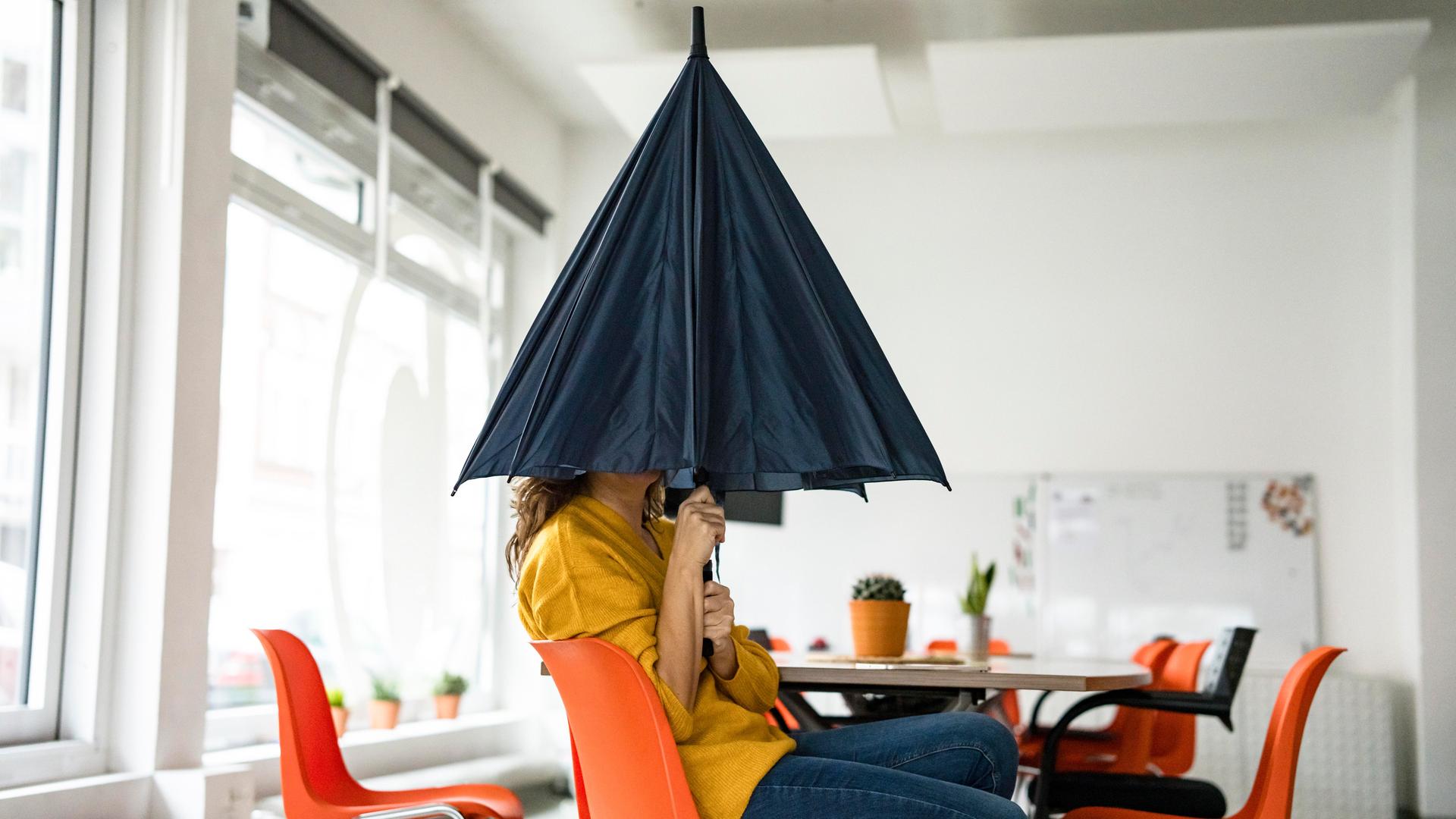 Eine Frau sitzt in einem Großraumbüro und versteckt ihren Kopf unter einem Regenschirm.