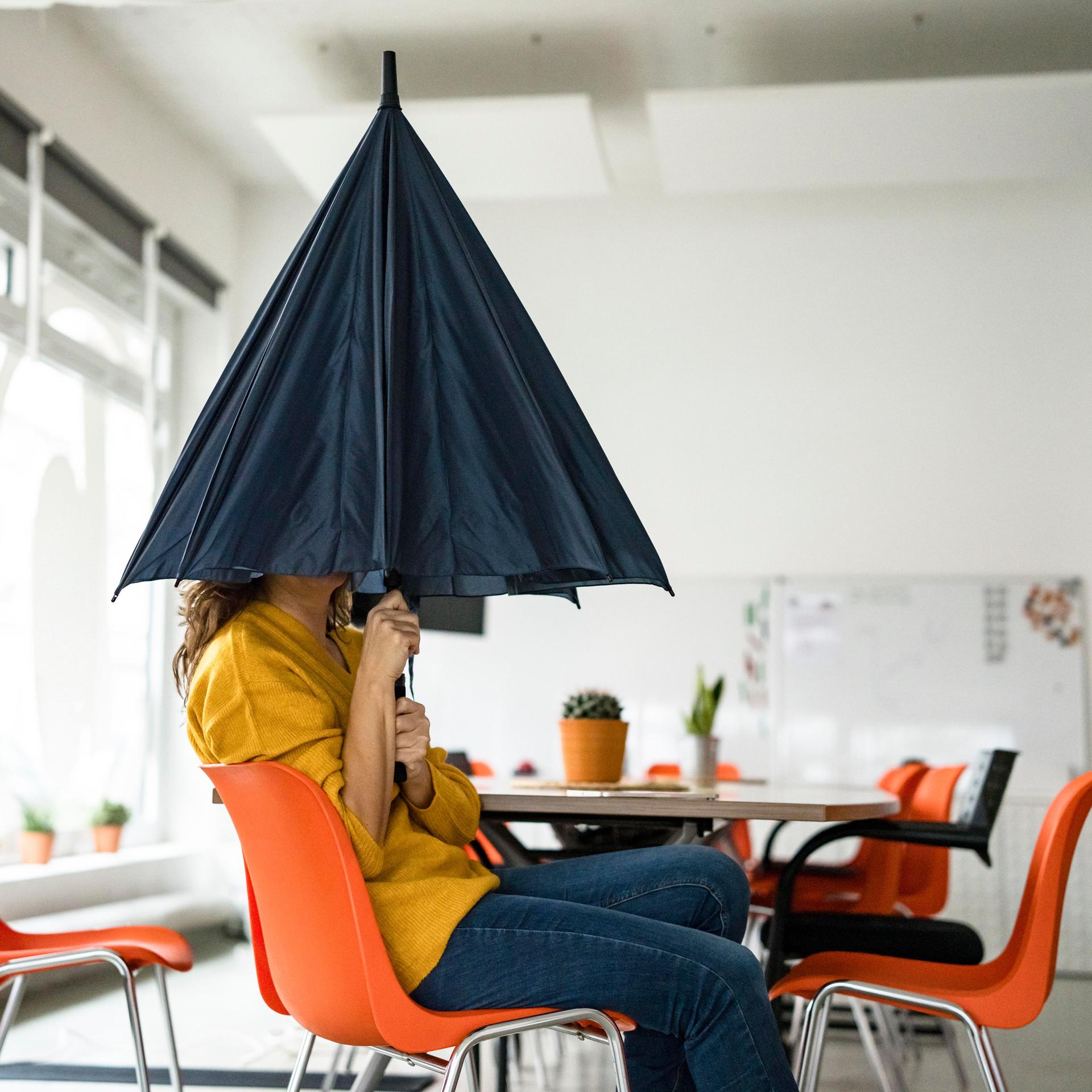 Eine Frau sitzt in einem Großraumbüro und versteckt ihren Kopf unter einem Regenschirm.