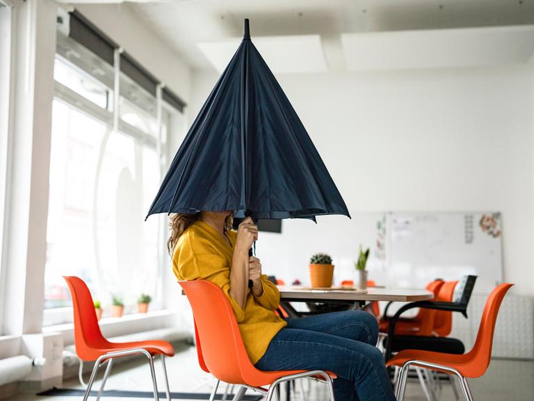 Eine Frau sitzt in einem Großraumbüro und versteckt ihren Kopf unter einem Regenschirm.