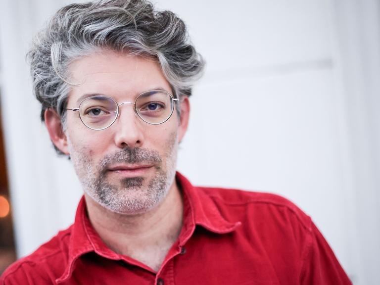 Der Autor Tomer Dotan-Dreyfus steht in rotem Hemd vor einer weißen Wand mit einem Fenster