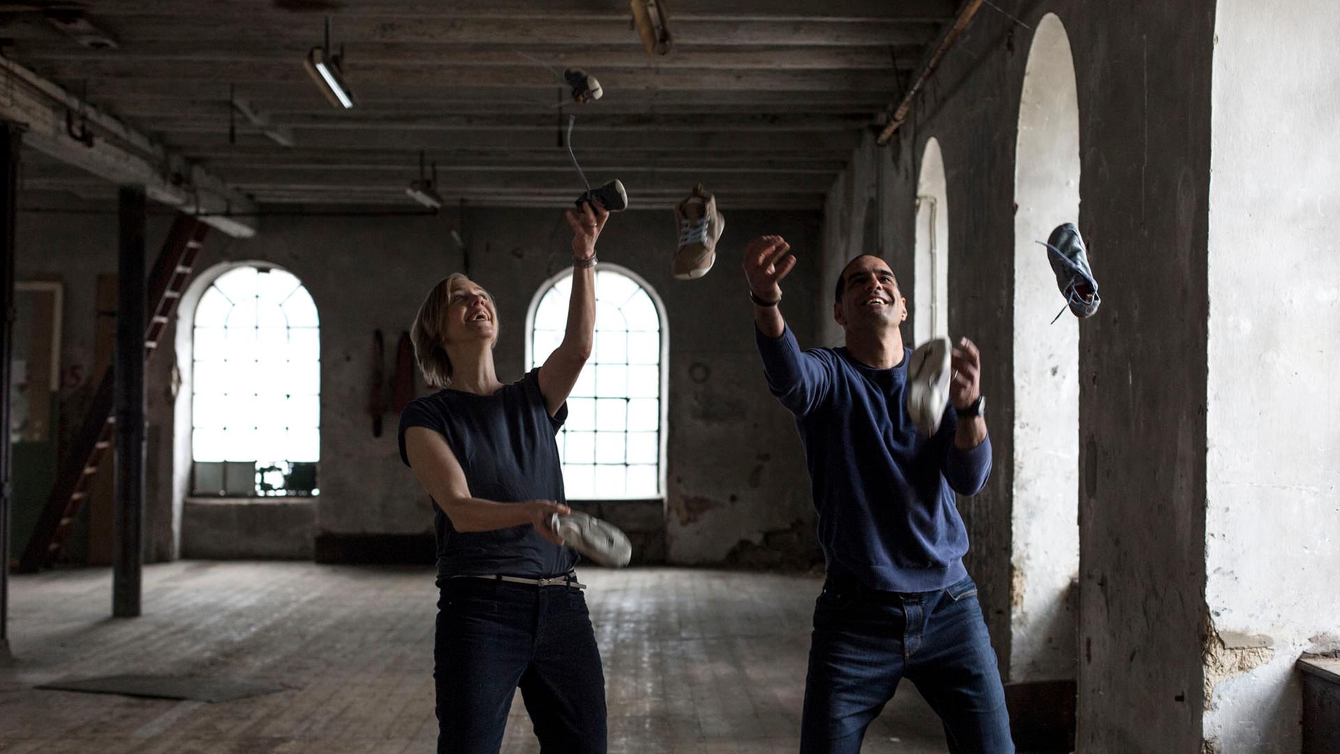 Anna und Ran Yona stehen in einem großen Raum und jonglieren mit Schuhen