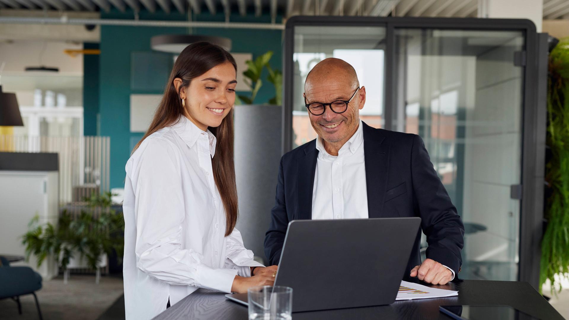 Das Foto zeigt eine Frau mit langen dunklen Haaren und einen Mann mit einem Laptop im Büro Das Foto zeigt eine Frau mit langen dunklen Haaren und einen Mann mit einem Laptop im Büro