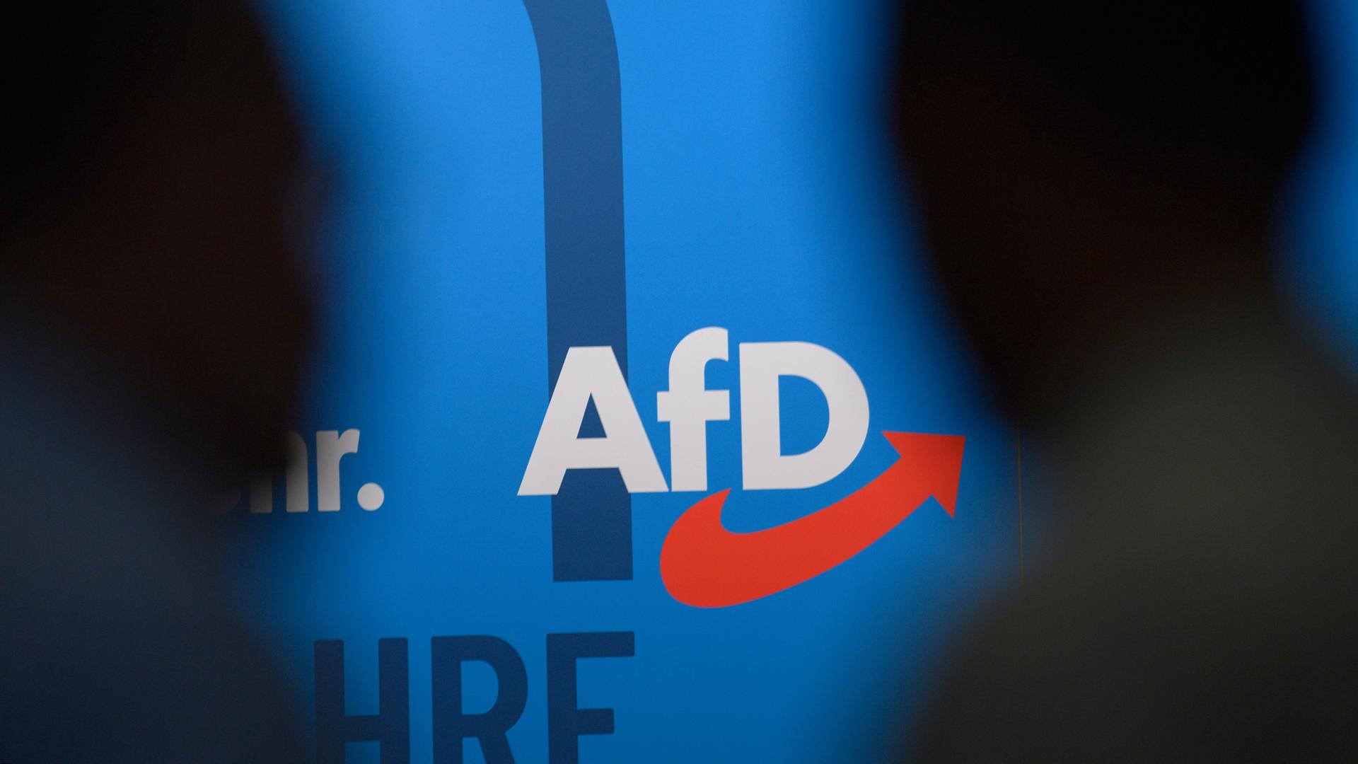 Auf einer blauen Wand steht das Logo der AfD, links und rechts die Silhouetten zweier dunkler Köpfe.