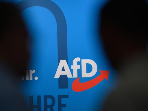 Auf einer blauen Wand steht das Logo der AfD, links und rechts die Silhouetten zweier dunkler Köpfe.