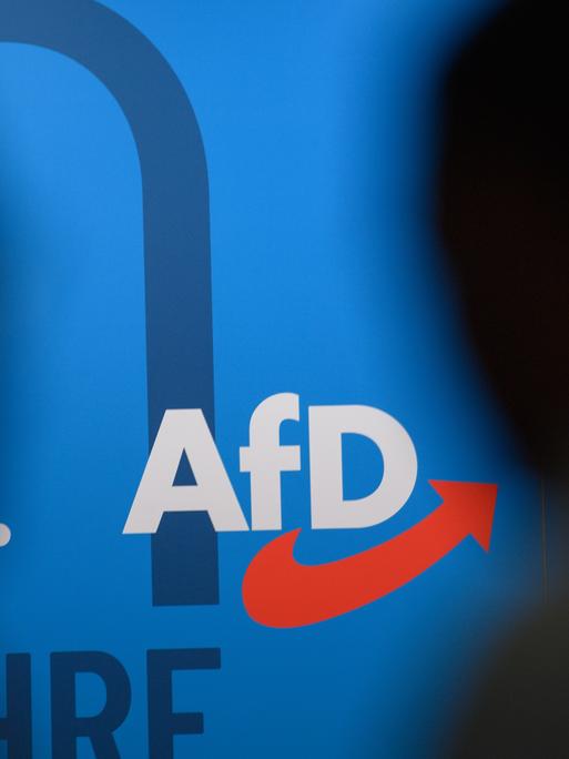 Auf einer blauen Wand steht das Logo der AfD, links und rechts die Silhouetten zweier dunkler Köpfe.