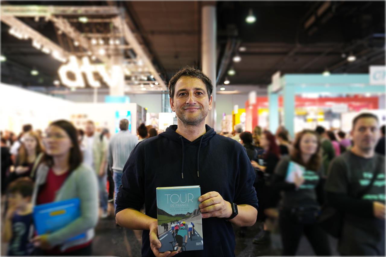 Stephan Klemm hält sein Buch "Tour de France" in der Hand und steht mitten in der Menschenmenge in der Messehalle der Frankfurter Buchmesse. 