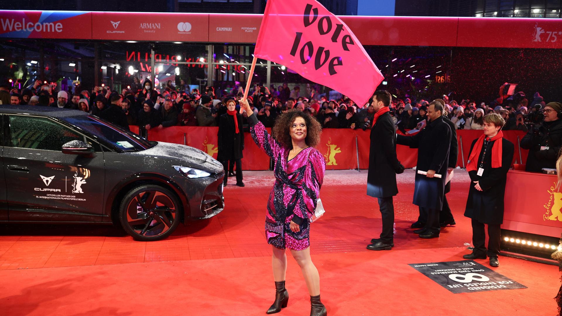 Schauspielerin Denise M'Baye steht mit einer pinken Flagge mit der Aufschrift "vote love" am Eröffnungsabend der Berlinale auf dem verschneiten Roten Teppich.