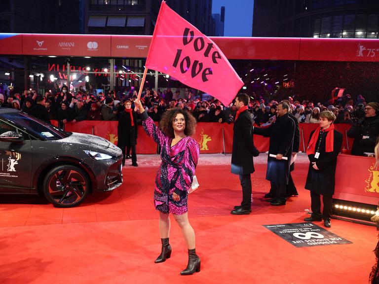 Schauspielerin Denise M'Baye steht mit einer pinken Flagge mit der Aufschrift "vote love" am Eröffnungsabend der Berlinale auf dem verschneiten Roten Teppich.