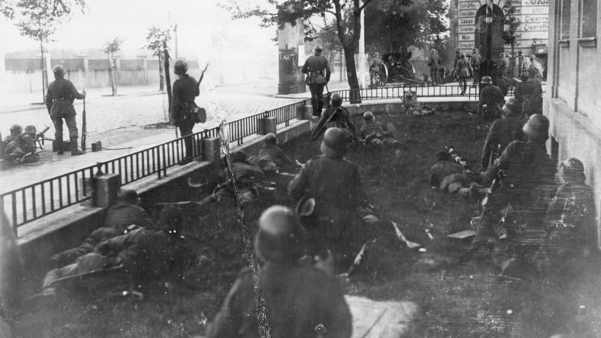 Schwarz-weiß Foto der Schwarzen Reichswehr beim Kampf um den Bahnhof in Myslowitz. Die Soldaten liegen in einer Art Vorgarten und stehen am Zaun darum. Schwarz-weiß Foto der Schwarzen Reichswehr beim Kampf um den Bahnhof in Myslowitz. Die Soldaten liegen in einer Art Vorgarten und stehen am Zaun darum.