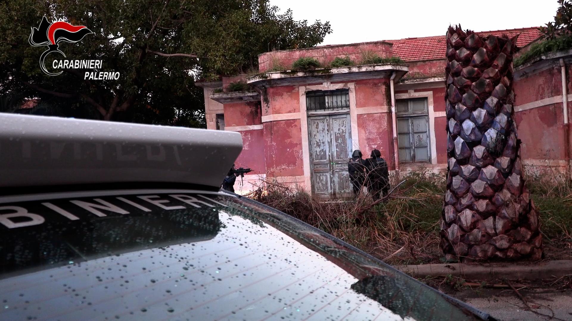 Blick über die Frontscheibe eines Polizeiautos auf ein baufälliges weinrotes Haus, vor dem Unkraut wuchert und eine abgebrochene Palme steht. Davor stehen zwei Polizisten, die als Silhouette zu sehen sind.