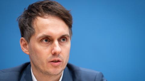 Ein Mann mit kurzen dunklen Haaren sitzt an einem langen Tisch und spricht.