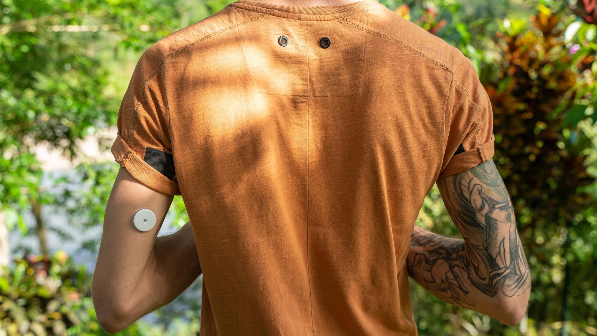 Ein Mann steht mit einem orangefarbenen T-Shirt in der Natur, am linken hinteren Oberarm trägt er einen Blutzucker-Sensor. Ein Mann steht mit einem orangefarbenen T-Shirt in der Natur, am linken hinteren Oberarm trägt er einen Blutzucker-Sensor.