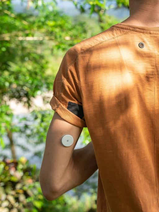 Ein Mann steht mit einem orangefarbenen T-Shirt in der Natur, am linken hinteren Oberarm trägt er einen Blutzucker-Sensor.