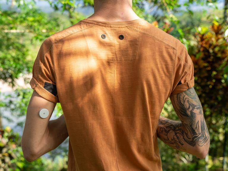 Ein Mann steht mit einem orangefarbenen T-Shirt in der Natur, am linken hinteren Oberarm trägt er einen Blutzucker-Sensor.