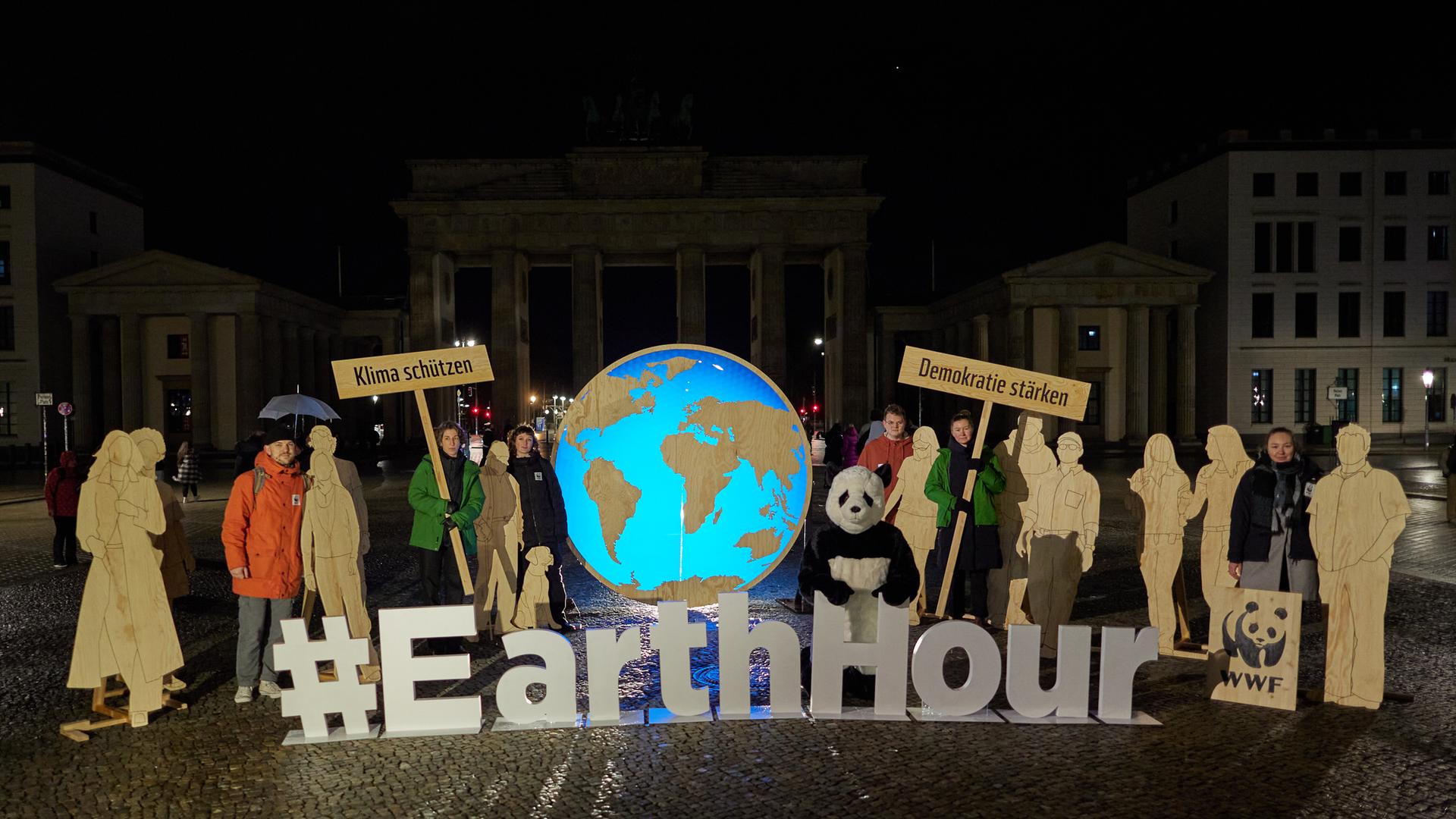 Im Hintergrund schemenhaft zu erkennen ist das Brandenburger Tor. Davor stehen Aktivisten mit Schildern, auf denen "Klima schützen" steht und "Demokratie stärken". Vor ihnen bilden aufgestellte Buchstaben den Begriff "Earth Hour".
