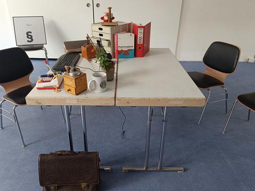 Bühnenbild aus dem Stück "Der Paragraphenreiter" vom "Theater der Verrückten" in Berlin Neukölln. Zu sehen ist ein voll gestellter Schreibtisch mit Aktenordner, Kaffemühle, Keyboard und weiteren amtstypischen Objekten