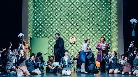 Vor einer großen Wand mit knalligem grünen Muster ist eine Gruppe von gestikulierenden Menschen versammelt, die erschreckt nach oben sehen, wie zwei kleine Fallschirme mit einem Stück Papier aufsteigen.