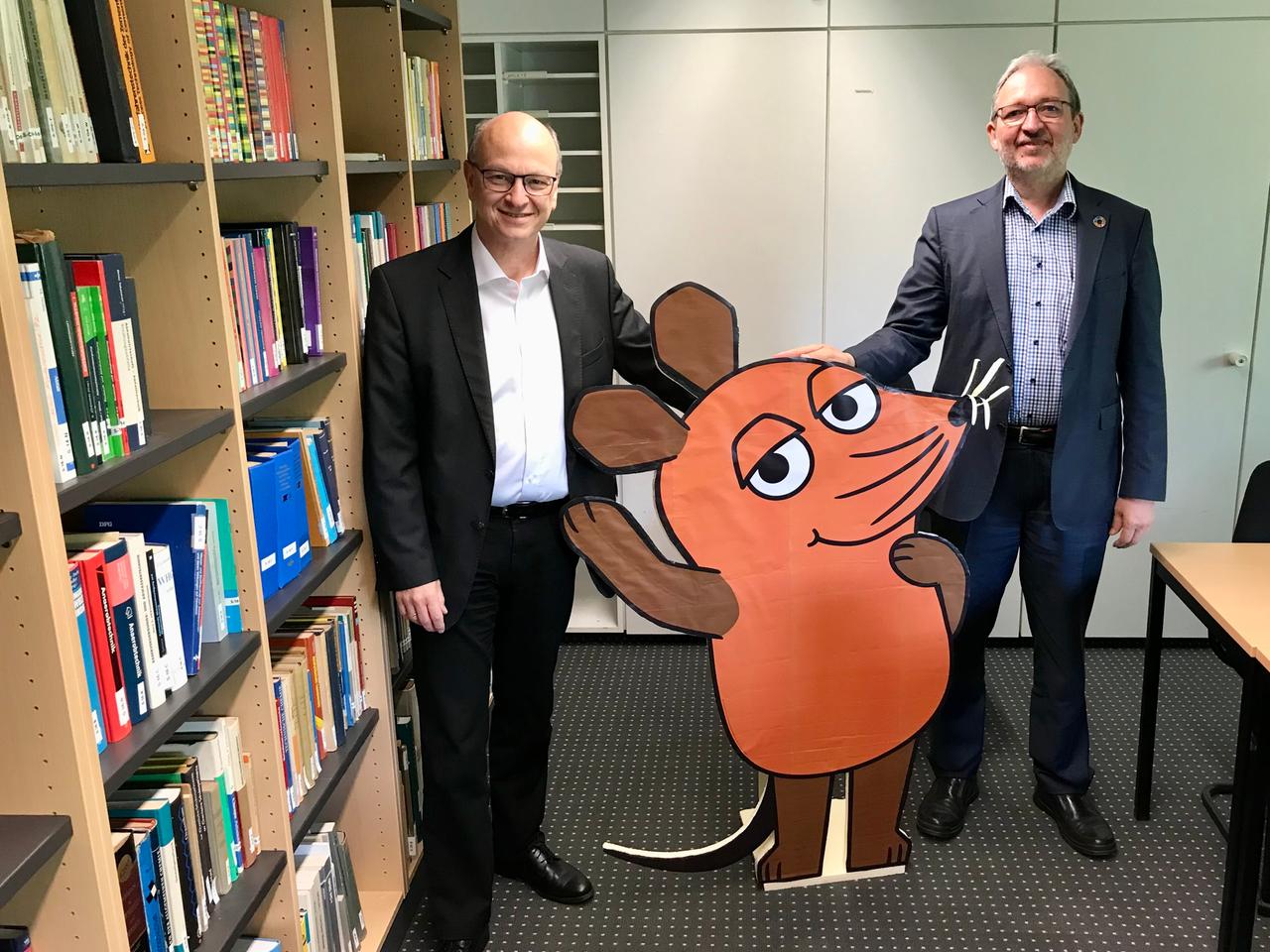 Daniel Goldmann und Dirk Schöps stehen in einem Innenraum. In ihrer Mitte haben sie eine zweidimensionale Mausfigur aus der ARD-Sendung "Die Sendung mit der Maus". Links sind Bücherregale zu sehen. Goldmann und Schöps tragen dunkle Zeugnisse. 