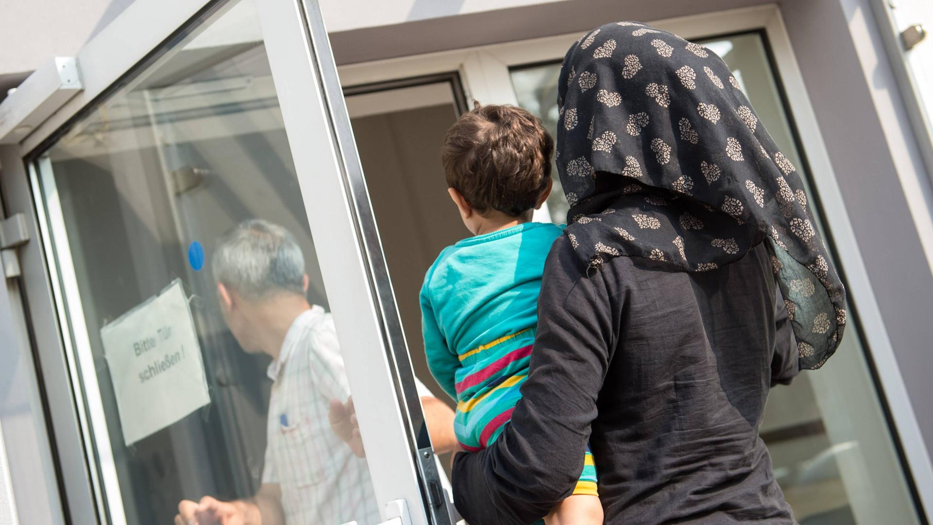 Eine verschleierte Frau mit einem Kleinkind auf dem linken Arm geht im August 2015 in Richtung einer geöffneten Tür, auf der ein Schild steht "Bitte Tür schließen". Die Tür gehört zur Ausländerbehoerde und BAMF-Außenstelle in Eisenhüttenstadt.