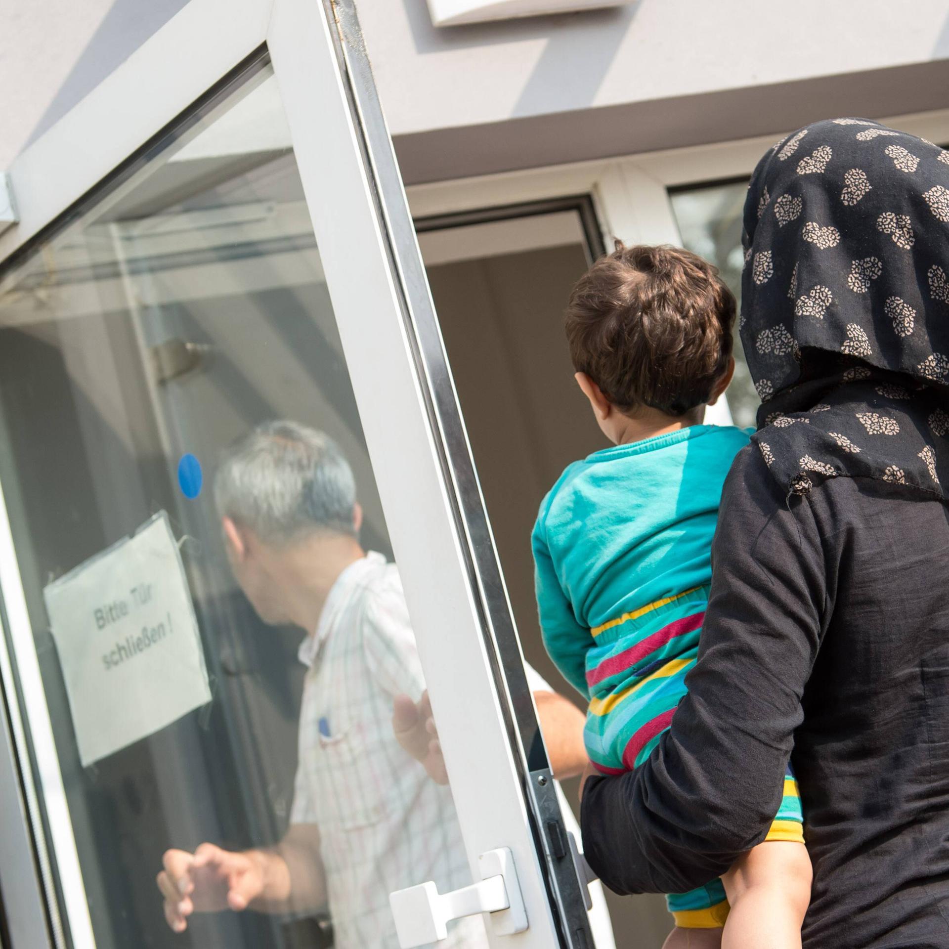 Eine verschleierte Frau mit einem Kleinkind auf dem linken Arm geht im August 2015 in Richtung einer geöffneten Tür, auf der ein Schild steht "Bitte Tür schließen". Die Tür gehört zur Ausländerbehoerde und BAMF-Außenstelle in Eisenhüttenstadt.