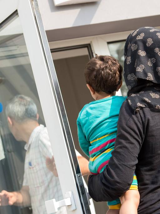 Eine verschleierte Frau mit einem Kleinkind auf dem linken Arm geht im August 2015 in Richtung einer geöffneten Tür, auf der ein Schild steht "Bitte Tür schließen". Die Tür gehört zur Ausländerbehoerde und BAMF-Außenstelle in Eisenhüttenstadt.