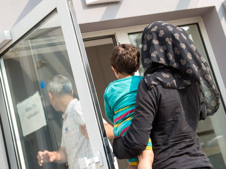 Eine verschleierte Frau mit einem Kleinkind auf dem linken Arm geht im August 2015 in Richtung einer geöffneten Tür, auf der ein Schild steht "Bitte Tür schließen". Die Tür gehört zur Ausländerbehoerde und BAMF-Außenstelle in Eisenhüttenstadt.