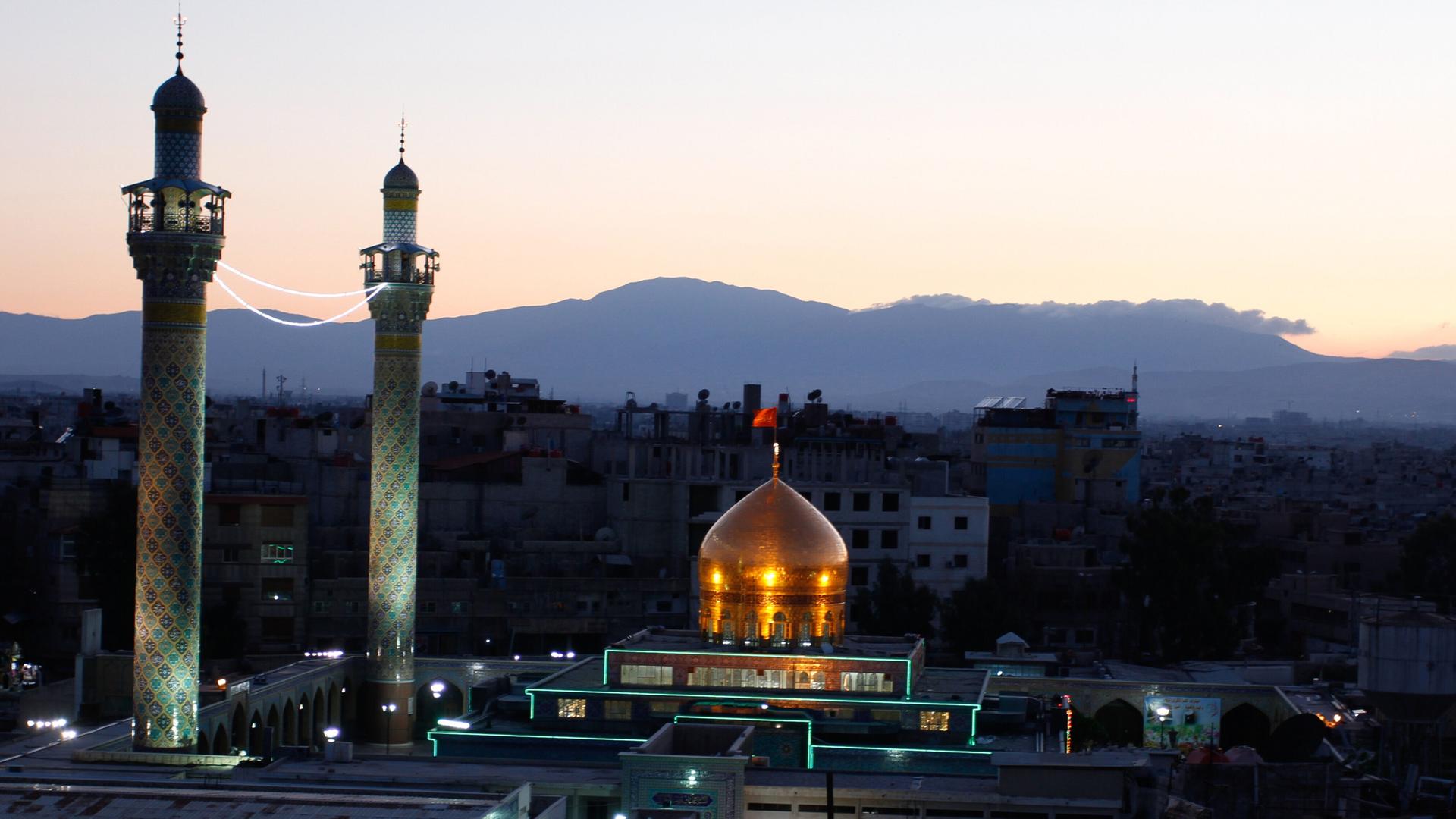 Das Mausoleum von Sajeda Seinab nahe Damaskus in der Abenddämmerung, zwei Minarette und eine goldene Kuppel sind zu sehen.