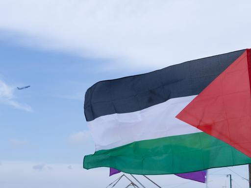 Die Palästinensische Flagge weht an einem Mast. Sie ist horizontal in den panarabischen Farben Schwarz, Weiß und Grün unterteilt. An der linken Seite ist ein rotes Dreieck.