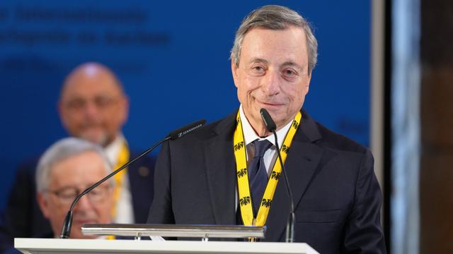 Mario Draghi spricht lächelnd in ein Mikrofon. Er trägt ein gelbes Band mit einer Medaille um den Hals.