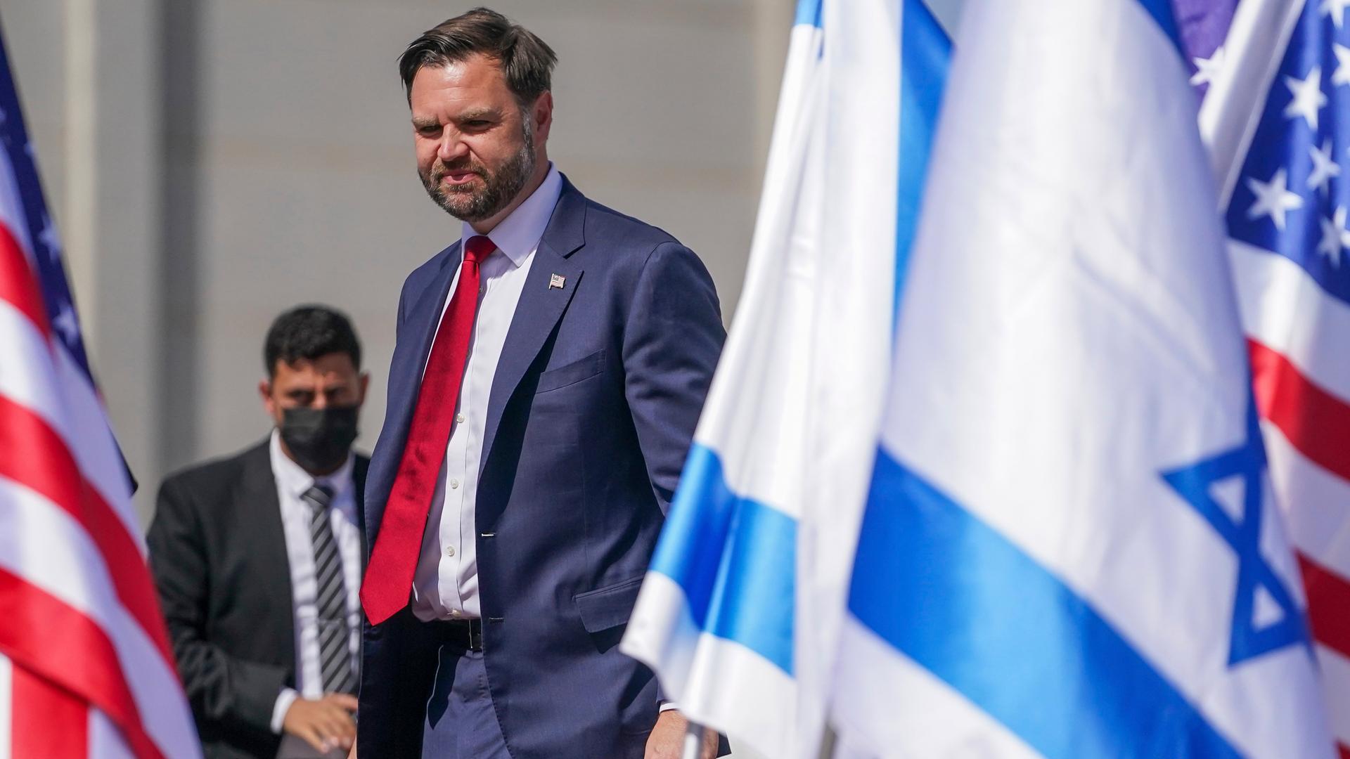 Vance im dunklen Jackett und mit roter Krawatte geht an auf dem Boden stehenden Flaggen Israels und der USA vorbei. Hinter ihm ein Security-Mann mit schwarzer Atemmaske.
