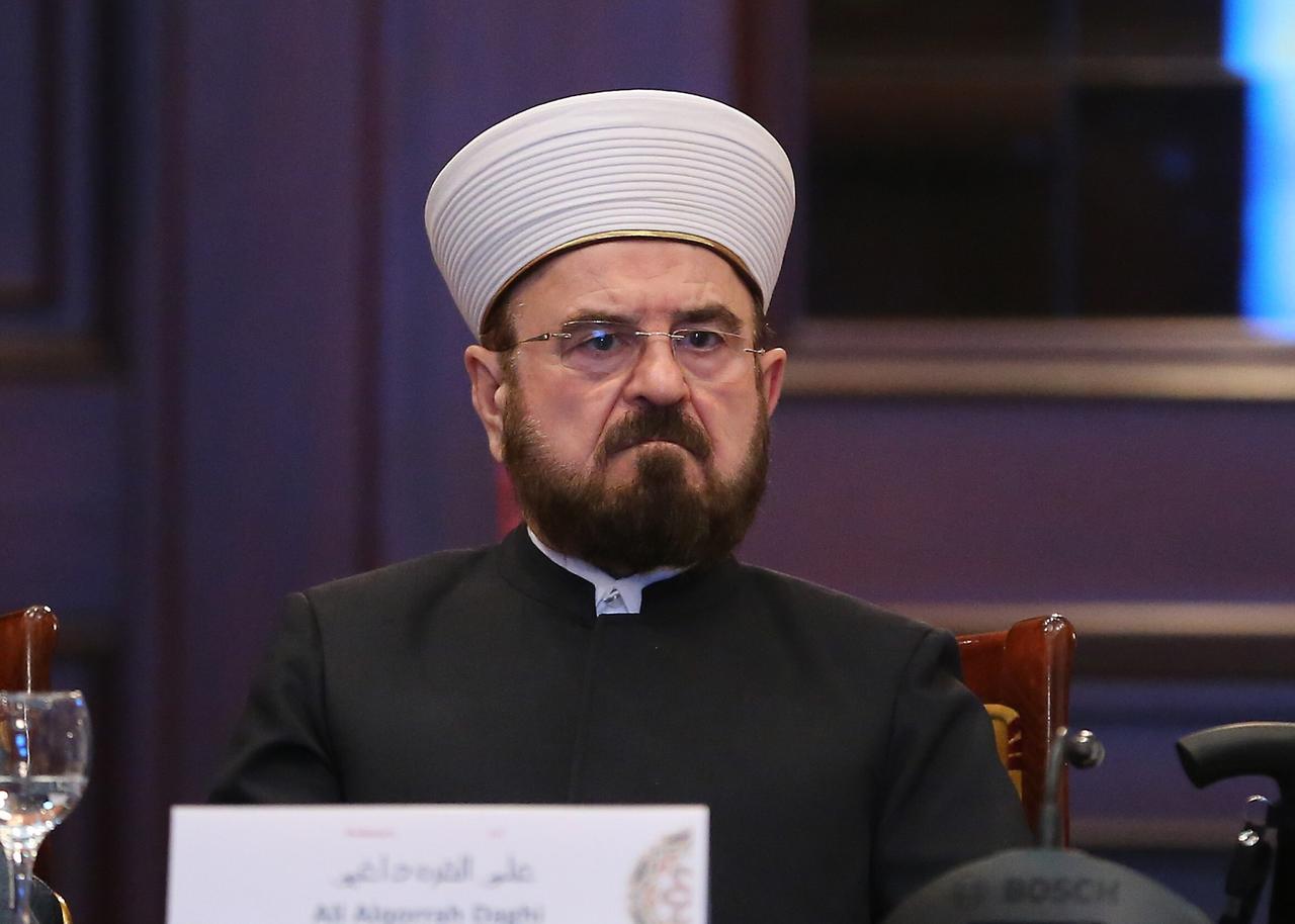 Ali al-Qaradaghi trägt dunkle Kleidung und eine weiße Mütze. Er hat dunkle Haare, trägt einen Vollbart und eine randlose Brille.